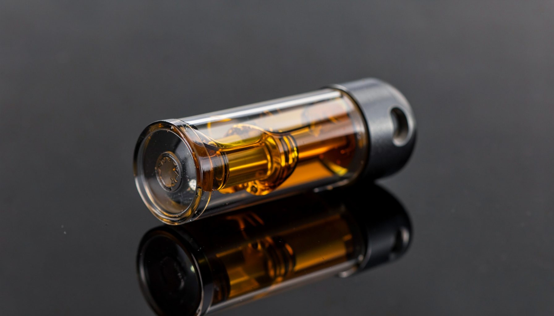 Close-up of a glass cylindrical container holding an amber-colored liquid, with a dark metal fitting, against a dark reflective surface