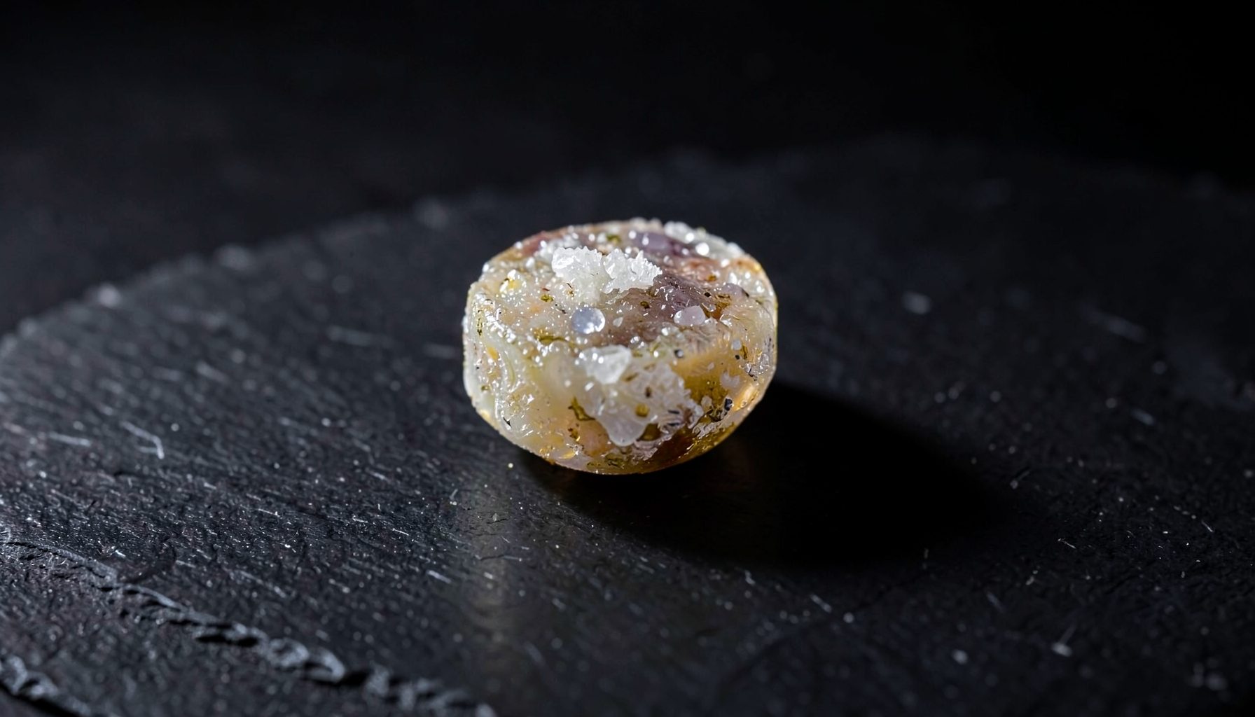 Small round crystalline substance with crystal formations on a dark textured surface