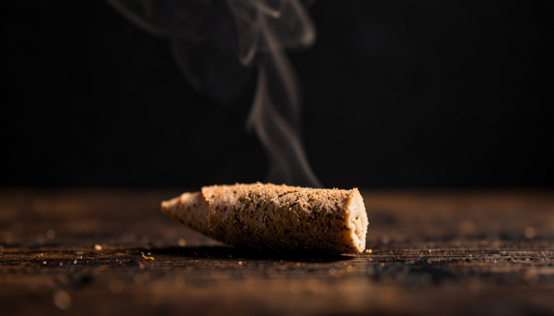 A close-up of a hand-rolled infused pre-roll resting on a dark wooden surface, with a soft amber glow and wisps of smoke