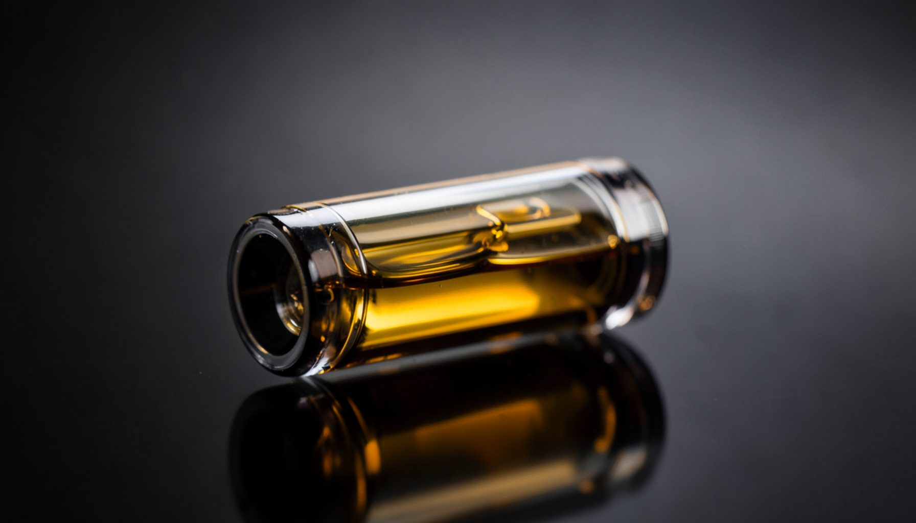 Vape cartridge with golden amber oil visible through the glass, placed on a dark reflective surface with soft gradient light