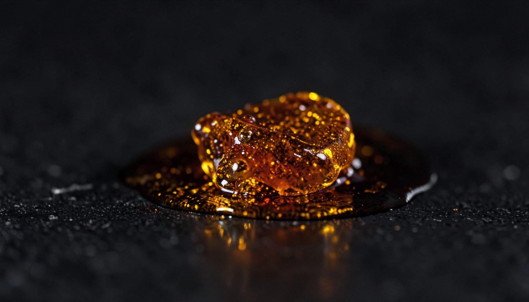 Macro shot of a glossy amber-colored concentrate blob on a dark textured surface, warm gold and brown tones